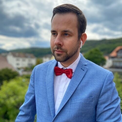 Lächelnde Person mit einer roten Mütze mit einem Hahnemblem, in legerer Kleidung vor einem unscharfen Hintergrund.